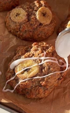 A spoon drizzling white icing over golden baked banana fritters topped with caramelised banana slices on parchment paper