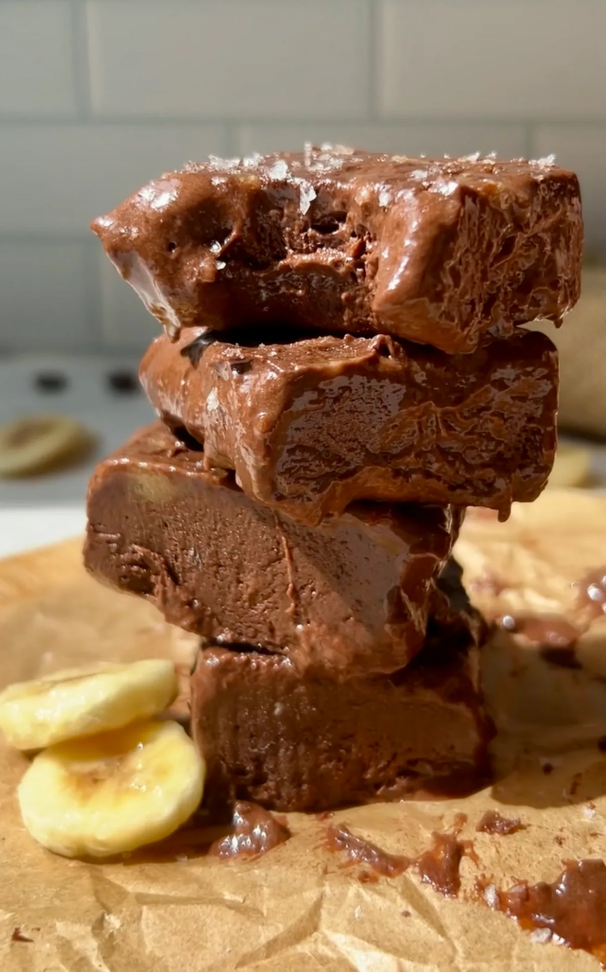 A stack of chocolate banana freezer fudge squares on parchment paper with banana slices scattered around and sea salt flakes on top against a white tile background