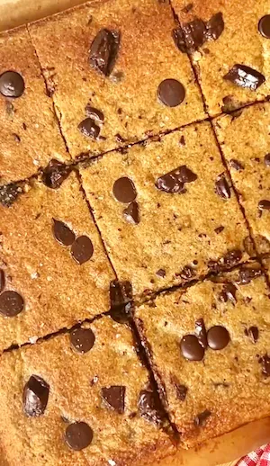 A hand holding a chocolate chip banana bread bar showing the golden fluffy texture with melted chocolate chips and sea salt on top with more bars visible in the background
