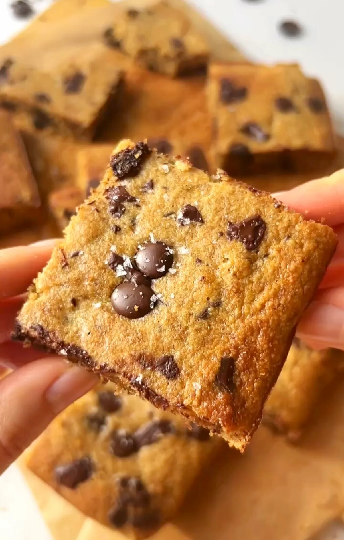 A hand holding a chocolate chip banana bread bar close up showing the golden fluffy texture with melted chocolate chips and sea salt flakes on top with more bars scattered in the background
