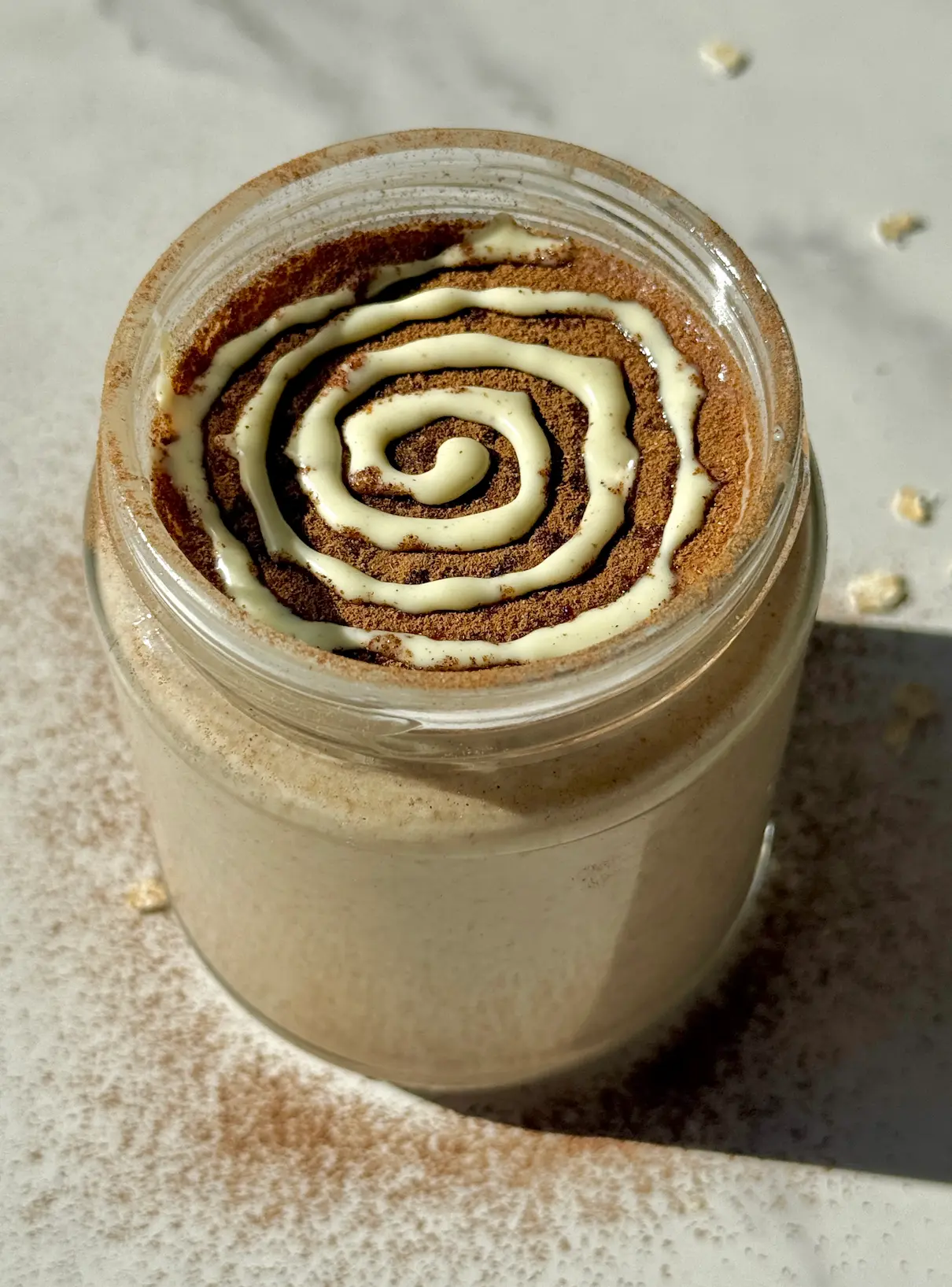 Blended cinnamon roll overnight oats in a jar with a white chocolate swirl on top and oats scattered around