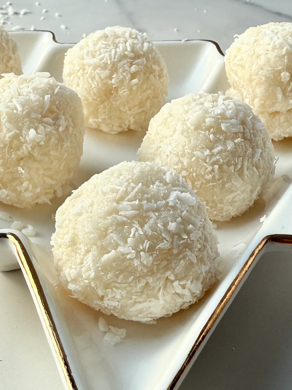 Healthy 3 ingredient coconut truffles rolled in shredded coconut arranged on a white and gold rimmed dish on a marble surface