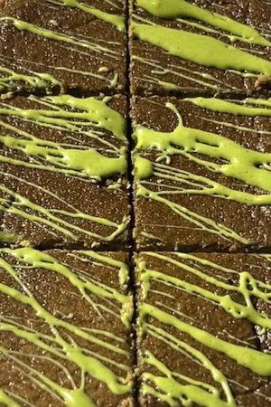 A hand holding a square slice of easy healthy matcha cake with bright green matcha icing drizzled on top and more cake slices on parchment paper behind