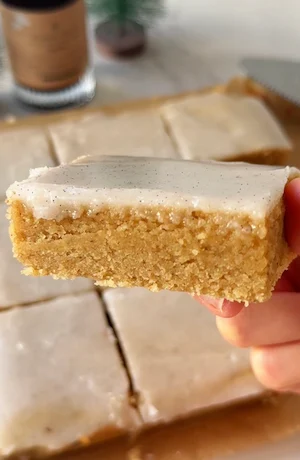 A hand holding a single gluten free vanilla white chocolate brownie square topped with vanilla bean frosting