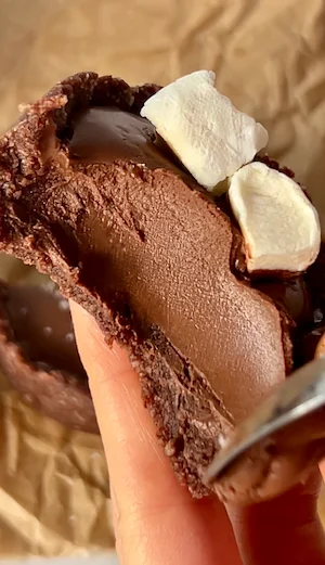 Two hot chocolate fudge cups on parchment paper with one broken open showing the rich glossy fudge filling inside the chocolate crust topped with white mini marshmallows