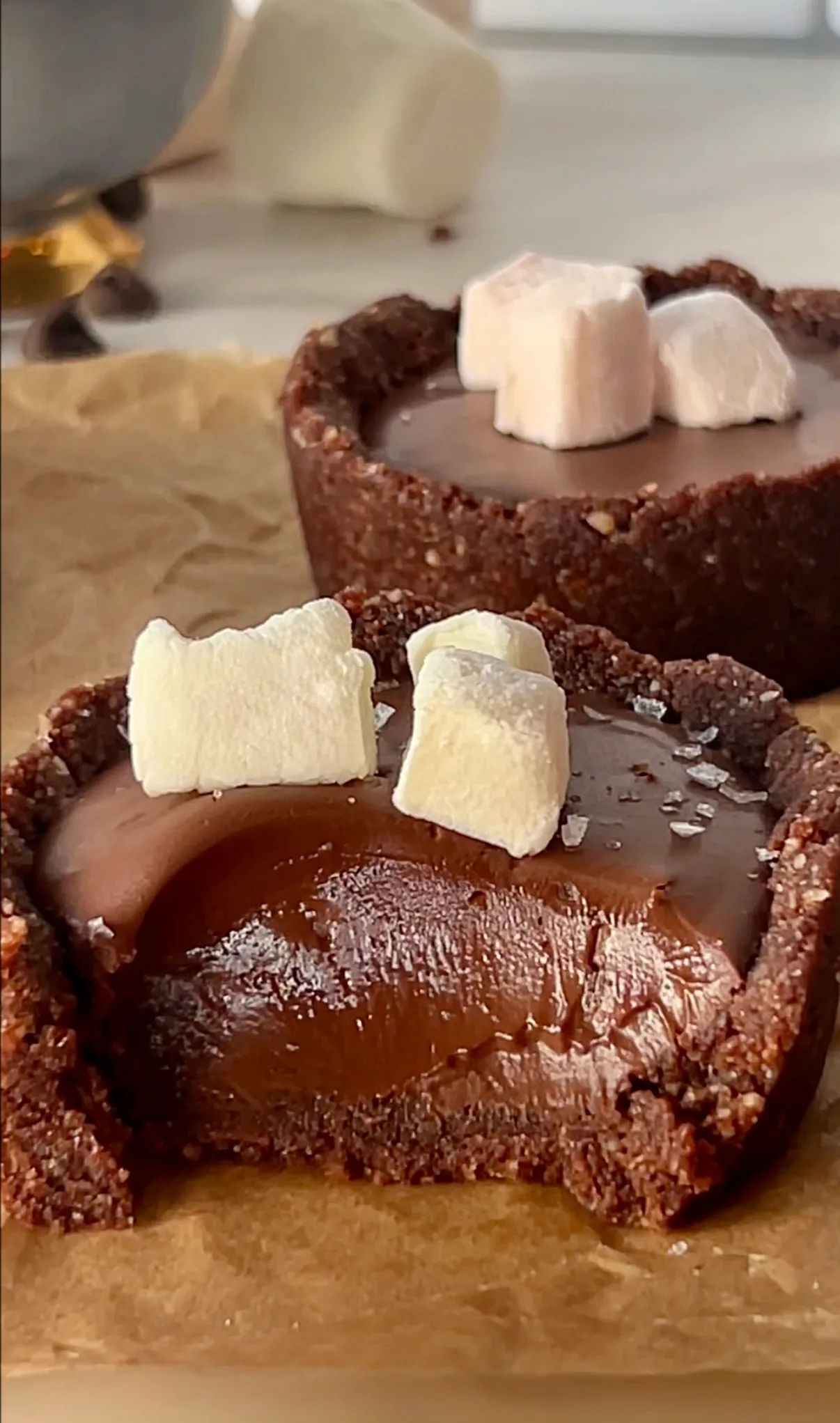 Two hot chocolate fudge cups on parchment paper with the front cup broken open showing the rich glossy fudge filling inside the dark chocolate crust topped with white mini marshmallows and sea salt flakes