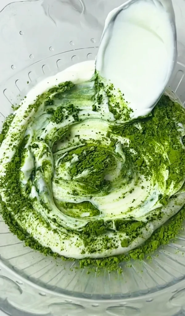 Matcha powder being folded into thick yogurt in a glass mixing bowl