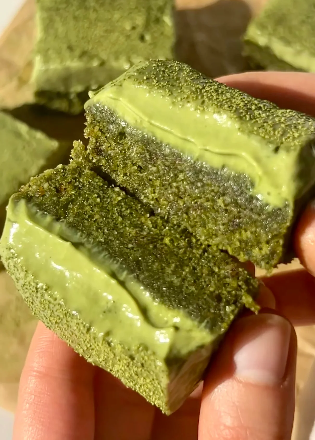 A hand holding a no bake matcha ganache bar broken in half revealing the dark green chewy cookie dough base and bright green white chocolate matcha ganache layer with more bars in the background on a wooden board