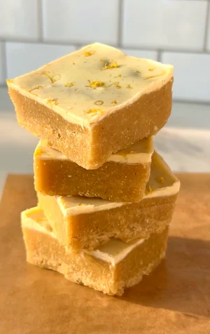 A stack of no-bake lemon bars with a creamy white chocolate lemon top and golden almond base, garnished with lemon zest on parchment paper