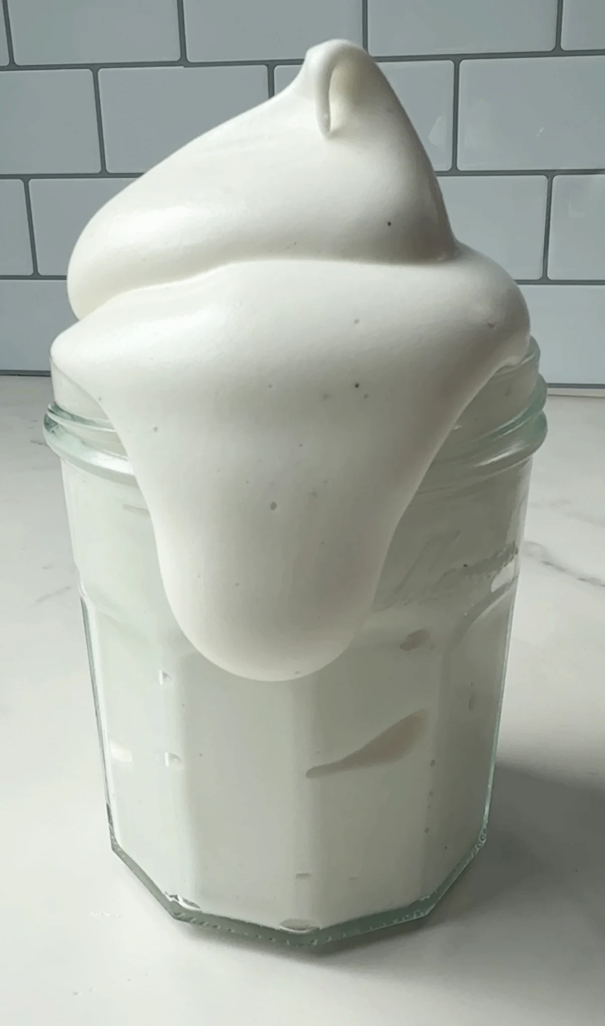 Vanilla bean marshmallow fluff piped into a tall swirl on top of a glass jar on a marble surface with a white tile background